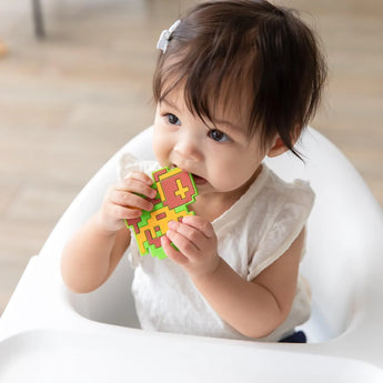 A toddler in a high chair, wearing a white shirt, explores the Bumkins Silicone Teether: Zelda™ Link shaped like cacti.