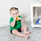 Baby sits on a gray carpet, smiling and holding a Bumkins Silicone Teether: Zelda™ Link, near a white shelf.