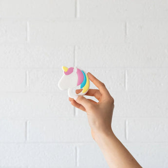 Hand holds Bumkins Silicone Teether: Unicorn, a non-toxic teether with vibrant details against a white brick wall.