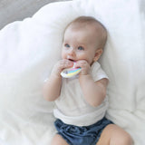 A baby in a white shirt and blue shorts enjoys a colorful Bumkins Silicone Teether: Unicorn on a white blanket.