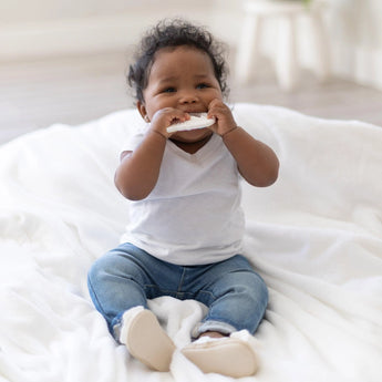 A baby in a white shirt and blue jeans happily chews on a Bumkins Silicone Teether: Unicorn while sitting on a soft blanket.