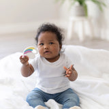 A baby on a white blanket holds a Bumkins Silicone Teether: Unicorn with a plant blurred in the background.