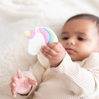 Baby in a beige outfit with Bumkins Silicone Teether: Unicorn on a soft, light-colored blanket.