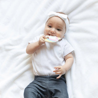 Baby in a headband and white onesie on a soft blanket, holding a colorful Bumkins Unicorn Silicone Teether.