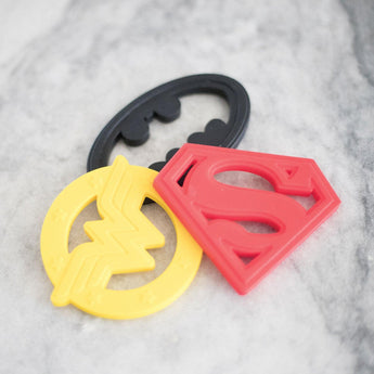 Three superhero silicone teethers by Bumkins: a red S shield, black bat silhouette, and yellow W for DC Comics fans.