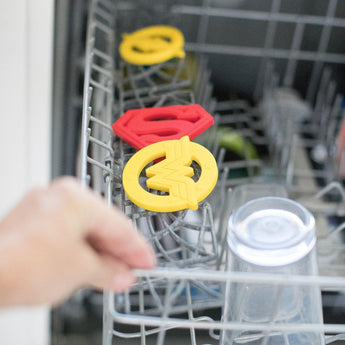 The Bumkins Silicone Teether: Wonder Woman rests on the top rack with glassware and dishes, showcasing its vibrant superhero theme.