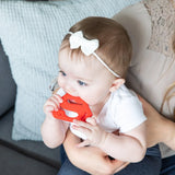 A baby in a white bow chews on a Bumkins Silicone Teether: Superman for gum relief, held by an adult on the sofa.