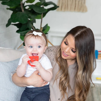 Woman holds a smiling baby chewing on a Bumkins Silicone Teether: Superman in a cozy room with a green plant framing the moment.