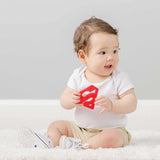 Baby in a white shirt and beige shorts holding a red Bumkins Superman Silicone Teether with curiosity on a soft carpet.