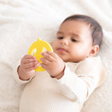 Baby holding a Bumkins Silicone Teether: Wonder Woman on a soft white blanket.