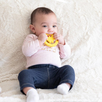 Baby on a fluffy blanket, in a pink sweater and blue pants, chewing on a yellow Bumkins Silicone Teether: Wonder Woman.