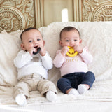 Two babies on a couch happily gnawing on Bumkins Silicone Teether: Wonder Woman. One wears a white shirt, the other a pink shirt.