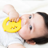 Baby on a white blanket, chewing a yellow Bumkins Wonder Woman silicone teether, wearing a light gray shirt.