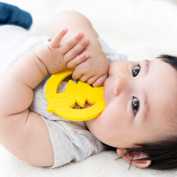 A baby chews on a yellow Bumkins Silicone Teether: Wonder Woman, wearing a light grey shirt.