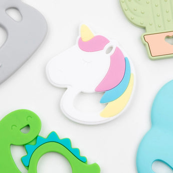 A vibrant Bumkins Unicorn Silicone Teether on a white surface, surrounded by textured dinosaur and cactus teethers.