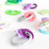 Bumkins Silicone Teething Charms in purple are scattered on a white surface, made from 100% food-safe silicone.