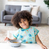 A curly-haired child smiles at the table, wearing a Bumkins Junior Bib: Ocean Life, with a bowl and spoon in front.