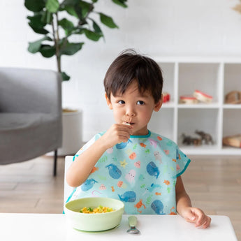 A child wearing a Bumkins Junior Bib: Ocean Life joyfully eats from a green bowl, with a shelf and plant softly framing the scene.