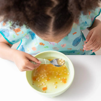 A curly-haired child sits at a table, eating soup from a light green bowl. Theyre holding a spoon and wearing Bumkins Junior Bib: Ocean Life, featuring colorful marine animals, waterproof fabric, and an adjustable fit. The soup contains small pasta and orange pieces in clear broth.