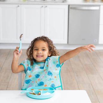 A curly-haired child sits at a table, holding a spoon and raising an arm excitedly. They wear a Bumkins Junior Bib: Ocean Life with adjustable fit, and have a blue plate of food in front of them. The kitchen is in the background.