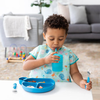 Child in a Bumkins Ocean Life Junior Bib, sipping from a blue cup and holding a fork at a table with sectioned food.