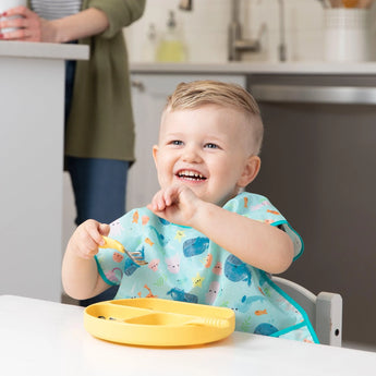 Child with short blond hair, happily eating with a spoon, wears a Bumkins Junior Bib: Ocean Life. Person in background.