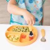 Child in a colorful Bumkins Ocean Life bib eating pasta, avocado, and fruit from an orange sectioned plate using an orange fork.