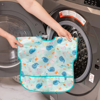 Person holding a Bumkins Junior Bib: Ocean Life with an adjustable fit in front of an open washing machine.