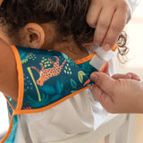 An adult adjusts a Bumkins Junior Bib: Jungle with waterproof fabric around a childs neck for an ideal fit.