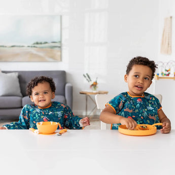 Two happy toddlers in matching outfits sit at a white table, wearing Bumkins Junior Bib: Jungle, eating from orange bowls.