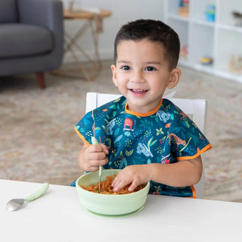 A child in a Bumkins Junior Bib: Jungle smiles, enjoying pasta from a green bowl at the table, thanks to its adjustable and waterproof design.