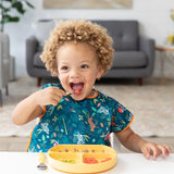 A curly-haired child wearing a Bumkins Junior Bib: Jungle happily eats from a divided yellow plate at the table with a sofa backdrop.