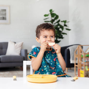 A child in a Bumkins Junior Bib: Jungle sits smiling at the table, holding melon, with a yellow plate and cutlery in front.