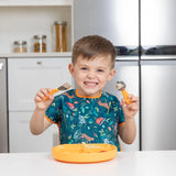 A child in a colorful Bumkins Junior Bib: Jungle smiles, holding a fork and spoon at the table beside a bowl.