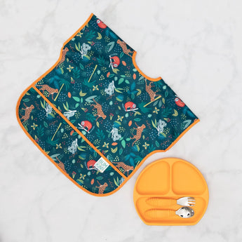 A Bumkins Junior Bib: Jungle with animal patterns pairs perfectly with a matching orange plate on a marble surface.