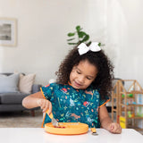 Young girl with curly hair in a bow and Bumkins Junior Bib: Jungle, eats indoors from an orange plate using a fork.