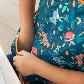 Child wears Bumkins Junior Bib: Jungle with colorful animals and leaves on a dark background, sitting at the table with hands together.