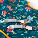 Gray baby utensils rest on a vibrant Bumkins Junior Bib: Jungle, adorned with animals and plants, featuring an adjustable fit.