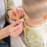 Person fastening a Bumkins Junior Bib: Camp Gear on a child, featuring colorful camping-themed illustrations.