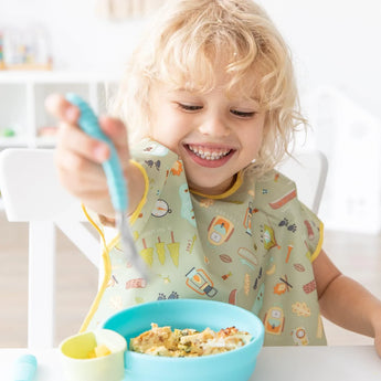 A curly blonde child enjoys a meal from a blue bowl, wearing a Bumkins Junior Bib: Camp Gear, seated playfully at the table.