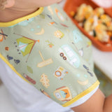 Child in a Bumkins Camp Gear Junior Bib with tents and trailers print, near food tray.