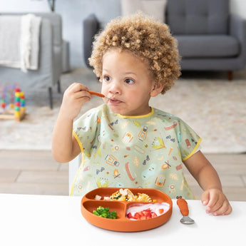 A curly-haired child eats from a divided plate, wearing a Bumkins Junior Bib in Camp Gear print. Cozy living room backdrop.