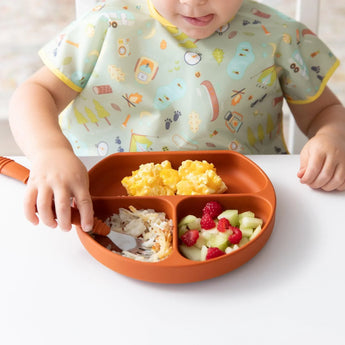 A child in a Bumkins Junior Bib: Camp Gear eats scrambled eggs, fruit, and more from a divided plate using a spoon.