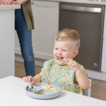 A smiling toddler wears a Bumkins Junior Bib: Camp Gear and eats from a divided plate as an adult stands nearby in the kitchen.