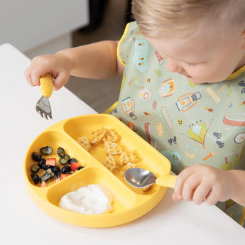 A toddler uses a fork/spoon to eat waffles, berries & yogurt from a yellow plate while wearing the colorful Bumkins Junior Bib: Camp Gear.