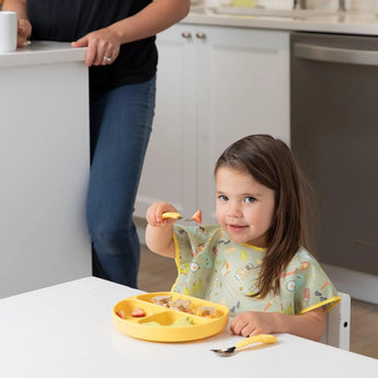 A girl in a Bumkins Junior Bib: Camp Gear eats from a yellow divided plate, with an adult nearby at the kitchen counter.