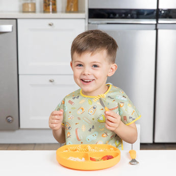 A smiling boy sits with a yellow plate, wearing a Bumkins Junior Bib: Camp Gear, holding a fork.