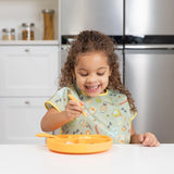 Child smiling while using a fork to eat from a yellow plate, wearing a colorful Bumkins Junior Bib: Camp Gear at the kitchen counter.