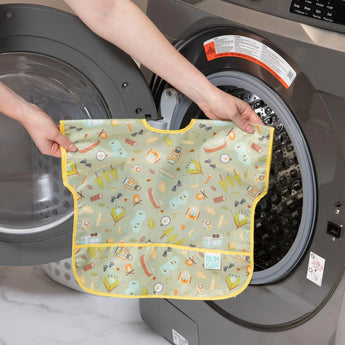 A person loads a Bumkins Junior Bib: Camp Gear into the washing machine.