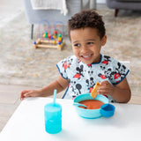 A child enjoys tomato soup and bread, wearing a Bumkins Junior Bib: Mickey Mouse Classic. Nearby, a blue sippy cup and toys sit.